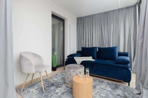 A bedroom styled with gray and blue tones, showcasing a plush bed with decorative pillows. Large windows let in natural light, complemented by airy curtains.