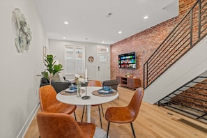 Interior view of dining room table and open living room area with beautiful exposed brick wall and 65" Roku smart TV.