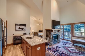 A modern open-plan kitchen and living area with a large island, stainless steel appliances, a stone fireplace, and floor-to-ceiling windows offering mountain views. A bedroom is partially visible.