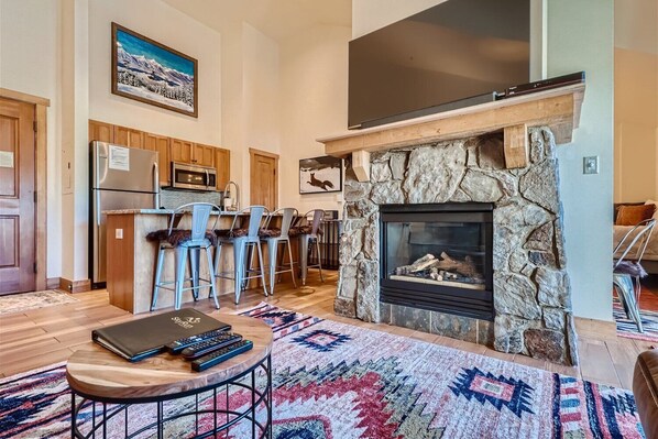 A cozy living space with a stone fireplace, flat-screen TV, and a wooden coffee table. The room features a modern kitchen with bar seating, colorful rugs, and mountain-themed wall art.