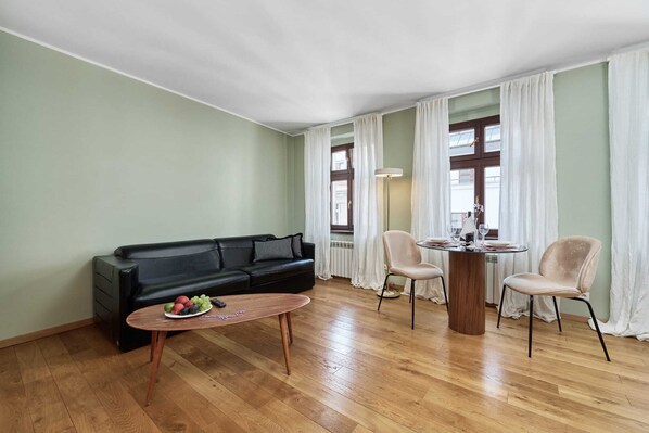 Spacious living room with a wooden floor, light curtains, and a black sofa. The room is bright and filled with natural light.