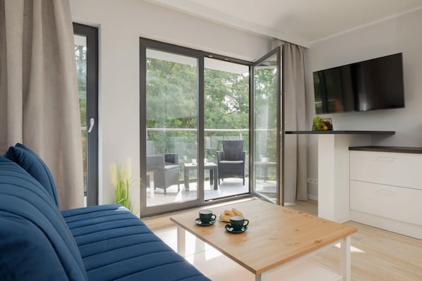 A cozy living room featuring a dark blue sofa, a light wooden coffee table, and large sliding glass doors opening to a balcony surrounded by lush greenery.