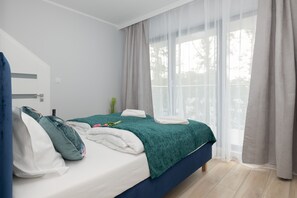  A neatly made bed with green bedding, white curtains letting in plenty of natural light, and minimalist decor.