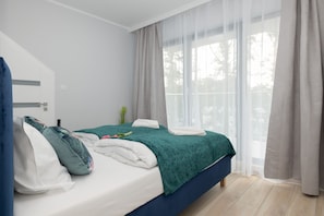 A neatly made bed with green bedding, white curtains letting in plenty of natural light, and minimalist decor.