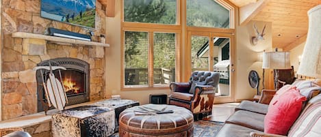 A cozy living room with a stone fireplace, wooden ceiling, large windows, a leather armchair, an ottoman, patterned rugs, and a TV mounted above the fireplace. Sunlight streams through the windows.