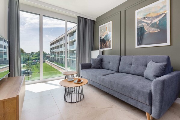 A bright living room featuring a comfortable grey sofa, stylish coffee table, and large windows with a view of lush greenery.
