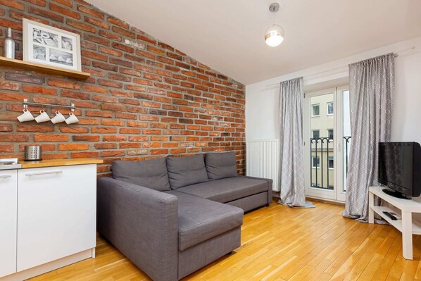 A cozy living area featuring a gray sofa, exposed brick walls, and bright wooden flooring, creating a warm and modern ambiance.
