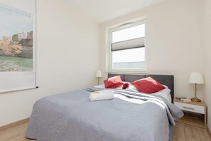 A bright bedroom featuring a double bed with gray bedding, red accents, and a minimalist design.