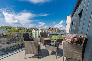 A balcony with comfortable outdoor seating, overlooking a scenic city view under a bright blue sky.