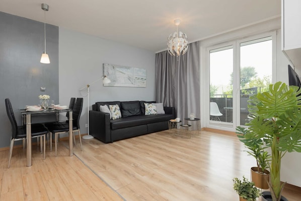 A bright living room with wooden flooring, a black leather sofa, a small dining table, and a large window.