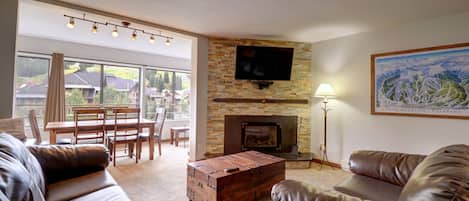 A cozy living room with leather sofas, a wooden trunk coffee table, a fireplace, and a wall-mounted TV. Large windows offer a view of an outdoor dining area with mountains in the background.