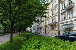 A charming city street with a classic building facade and lush green trees. Ornate balconies and tall windows lend a historic feel to the architecture.