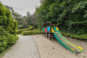 Outdoor playground with a slide and a pathway, surrounded by bushes and trees.