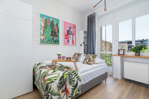 A bright, airy bedroom with colorful decor, a white bed, and large windows letting in natural light.
