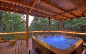 Covered deck with a hot tub and seating, surrounded by lush greenery.