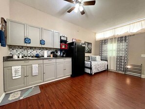 Refrigerator, microwave, toaster oven, coffee maker, cooking utensils, storage cabinets, sofa, exterior window with shade, ceiling fan