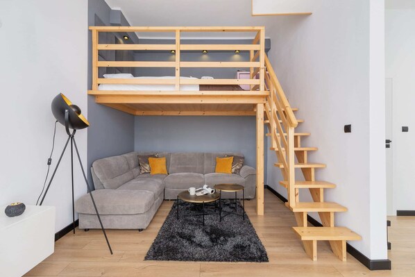 A living room with a loft-style bed, wooden stairs, a gray sofa, and a modern tripod lamp.
