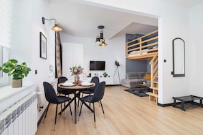 A spacious living room with a loft bed, gray sofa, and wooden accents, lit by modern lighting.
