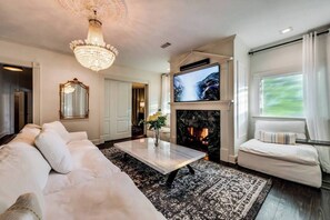 A spacious living room featuring a 55" Smart TV with a soundbar, complemented by an exquisite crystal chandelier and the inviting warmth of a wood-burning fireplace, creating an atmosphere of both modern comfort and timeless elegance.