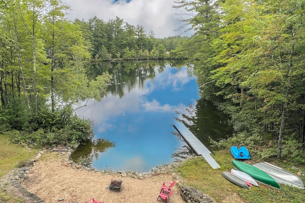 A long dock at a private, sandy beach on a New Hampshire lake is seen with kayaks on the ground and lots of trees surrounding the water.