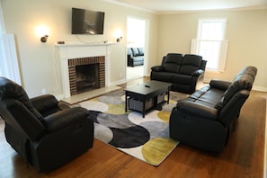 Living Room from Foyer showing Roku TV, wall mounted lamps, fireplace, reclinable single seat, loveset, and sofa, coffee table, Den in the background