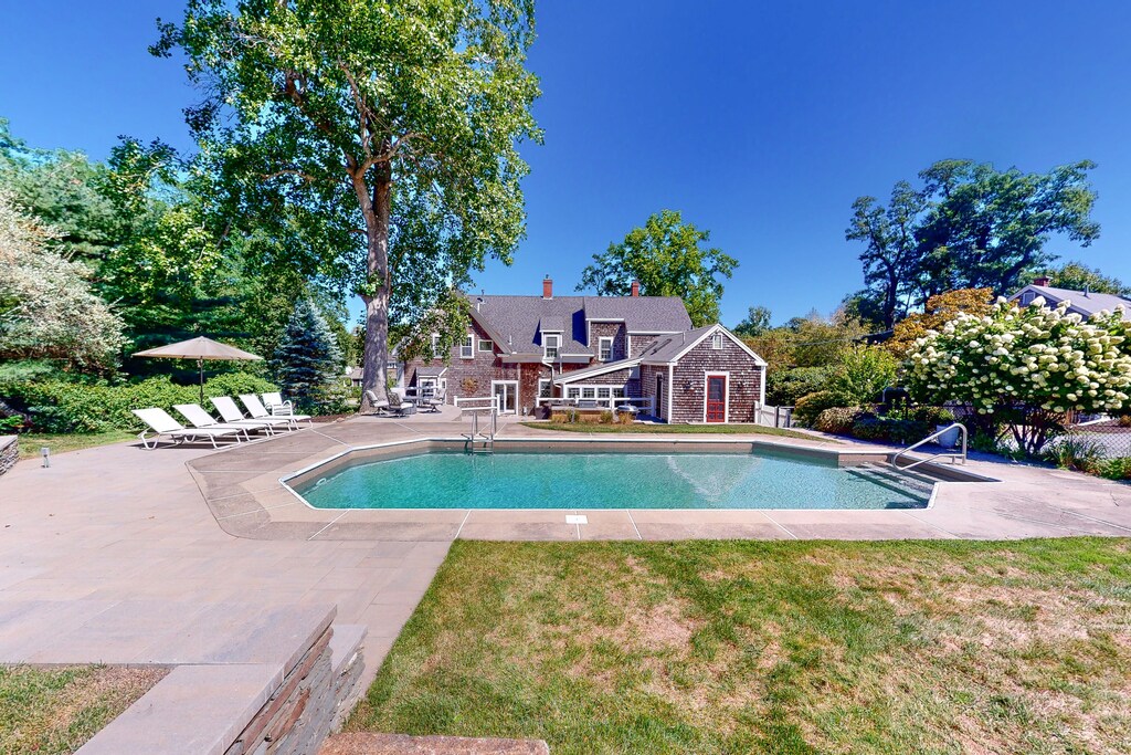 An inground pool at a Cape Cod vacation rental is seen in summer with green grass and lush trees surrounding