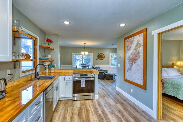 Explore our farmhouse-style kitchen with butcher block countertops and open shelving. Admire the Kodiak Island map as you cook on the stainless steel oven. View extends into bedroom and dining room, adding to the charm.
