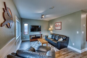 Glimpse into our living room adorned with black leather couches, a wall-mounted TV, and captivating artwork by local Kodiak artists. The pieces, featuring whales and Native Americans on horses, add cultural flair to the space.