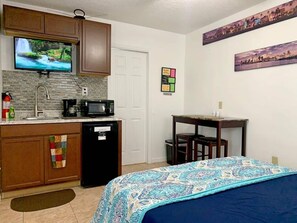 room with kitchenette, kitchen cabinets, microwave, coffee maker, small refrigerator, fire extinguisher, tv, dining table, decorations, access door to bathroom