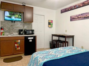 room with kitchenette, kitchen cabinets, microwave, coffee maker, small refrigerator, fire extinguisher, tv, dining table, decorations, access door to bathroom