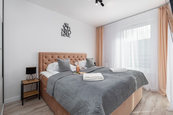 A cozy bedroom with a stylish bed, white and grey tones, and large windows allowing natural light.
