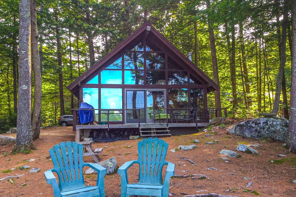 A lakehouse vacation rental in New Hampshire with a wall made entirely of windows sits surrounded by woods with chairs in front.
