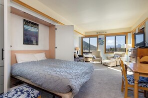 A well-lit studio apartment features a Murphy bed, chairs, a desk, and panoramic mountain views through large windows. A "Colorado" poster hangs above the bed.