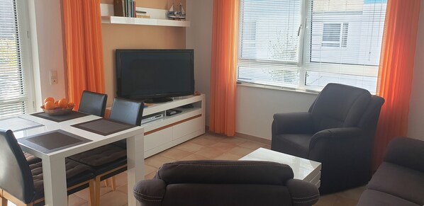 Wohnzimmer mit Sofa, zwei Sesseln, Esstisch und Bestuhlung sowie Sideboard und TV