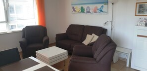 Wohnzimmer mit Sofa, zwei Sesseln, Esstisch und Bestuhlung sowie Sideboard und TV