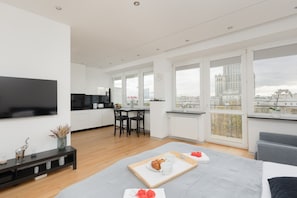 A bright open-plan living space with a sofa, dining area, and large windows overlooking the city.
