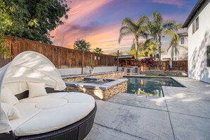 This view of our backyard highlights the heated pool and spa, surrounded by lush palm trees and a cozy lounge area. Relax on the sunbed, unwind by the water, or enjoy the peaceful ambiance under the beautiful sunset sky.