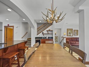 A rustic dining area features wooden furniture, a large antler chandelier, and an adjacent living space with a leather couch and framed artworks on the wall.