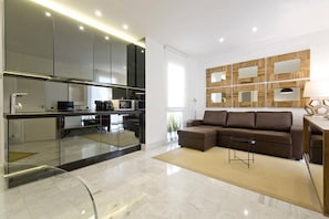 View of the living room, with a sofa,a television and design decoration. Next to the living room is the kitchen, fully equipped with ceramic hob, toaster, coffee maker and kitchen utensils.