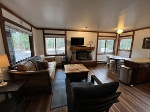 The living room has seating for four with a couch and chair situated around the wood fireplace located in a stone hearth, a 40” smart TV, and a dining area with seating for four tucked into a light-filled corner.