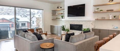 The living room is set up with everything you need for a relaxing space with an electrical fireplace, 65” smart TV, two sofas and two chairs, and well-placed decor creating a cozy home ambiance.