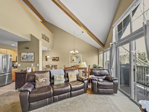 A spacious, well-lit living room with high ceilings, large windows, a leather sofa and recliner, a dining area in the background, and a view of trees outside.