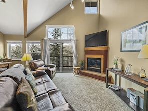 A cozy living room with leather sofas, a fireplace, a wall-mounted TV, and large windows overlooking a balcony with outdoor trees. The room features neutral tones and various decor pieces.