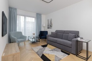 A bright living room with a gray sofa, modern carpet, and a small coffee table. A large window allows natural light to fill the space.