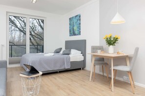 The sleeping area includes a comfortable double bed with gray bedding and a large window providing natural light and a view of the greenery outside.