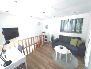 Wohnzimmer mit 2-Sitzer-Sofa, Sessel, Fernseher und Arbeitsplatz