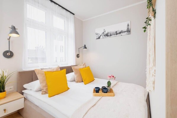 A bright bedroom with a double bed, vibrant yellow cushions, minimalist decor, and greenery accents, creating a cheerful and cozy sleeping space.
