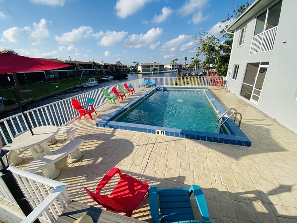 Pool overlooking canal with lots of seating, table with umbrella and more.