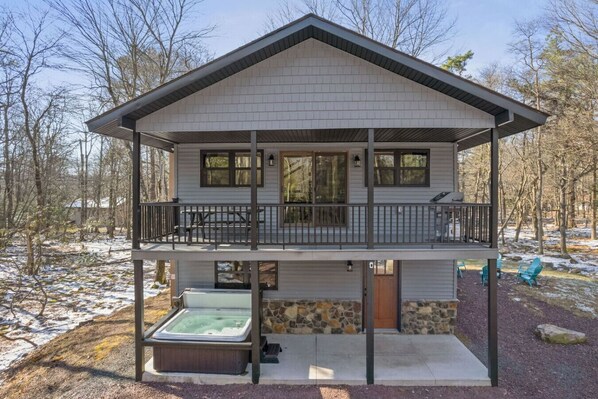 Front of House with a covered top deck and lower patio with a hot tub.