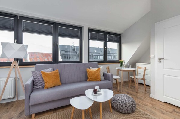 A bright and modern living room with a large gray sofa, decorative pillows in mustard yellow, and a round white coffee table. Natural light enters through large windows.